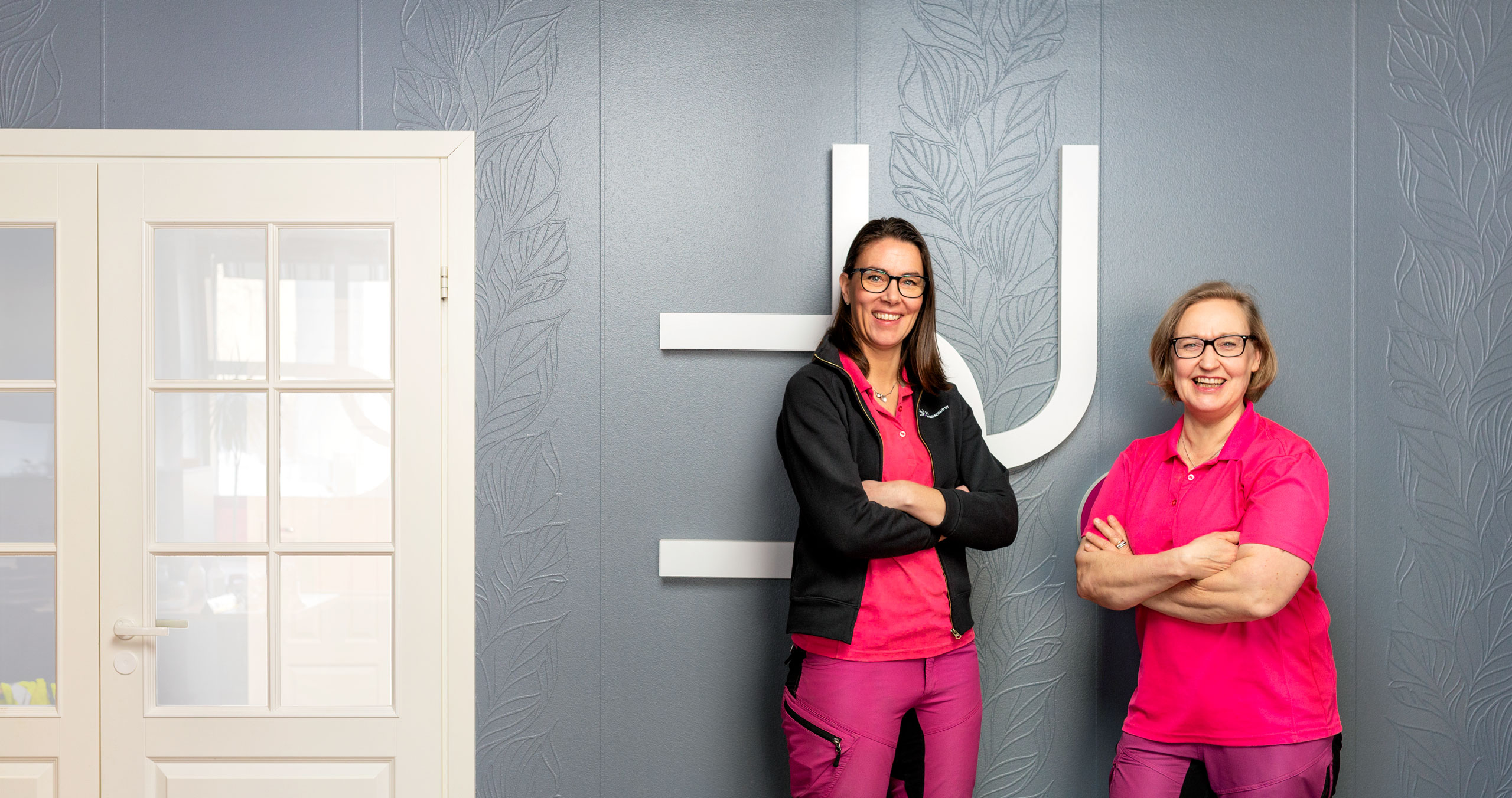 The founders of Duo Siivouspalvelu Oy, Mari Männistö and Sanna Kananen, stand in front of a beautiful gray wall with a white door.