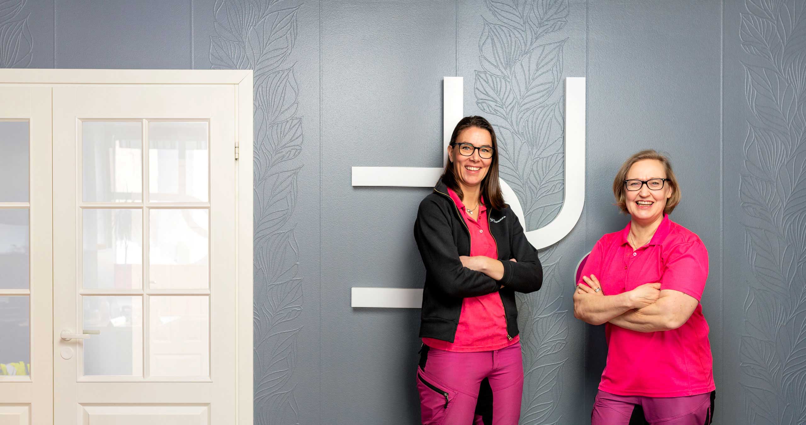 The founders of Duo Siivouspalvelu Oy, Mari Männistö and Sanna Kananen, stand in front of a beautiful gray wall with a white door.