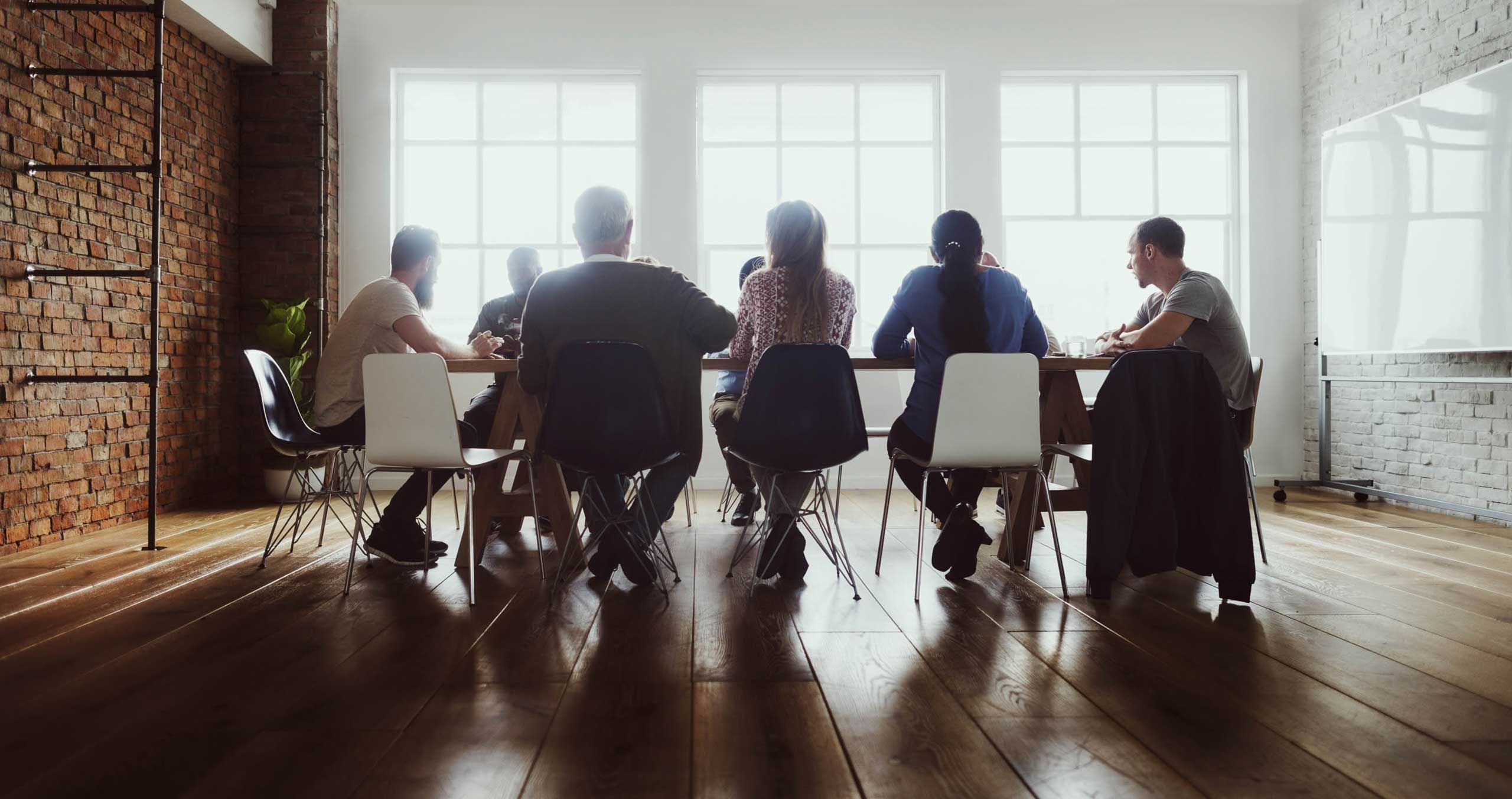 A group of people are sitting in a room at a conference table. There is a brown brick wall on the left and large checkered windows at the back.